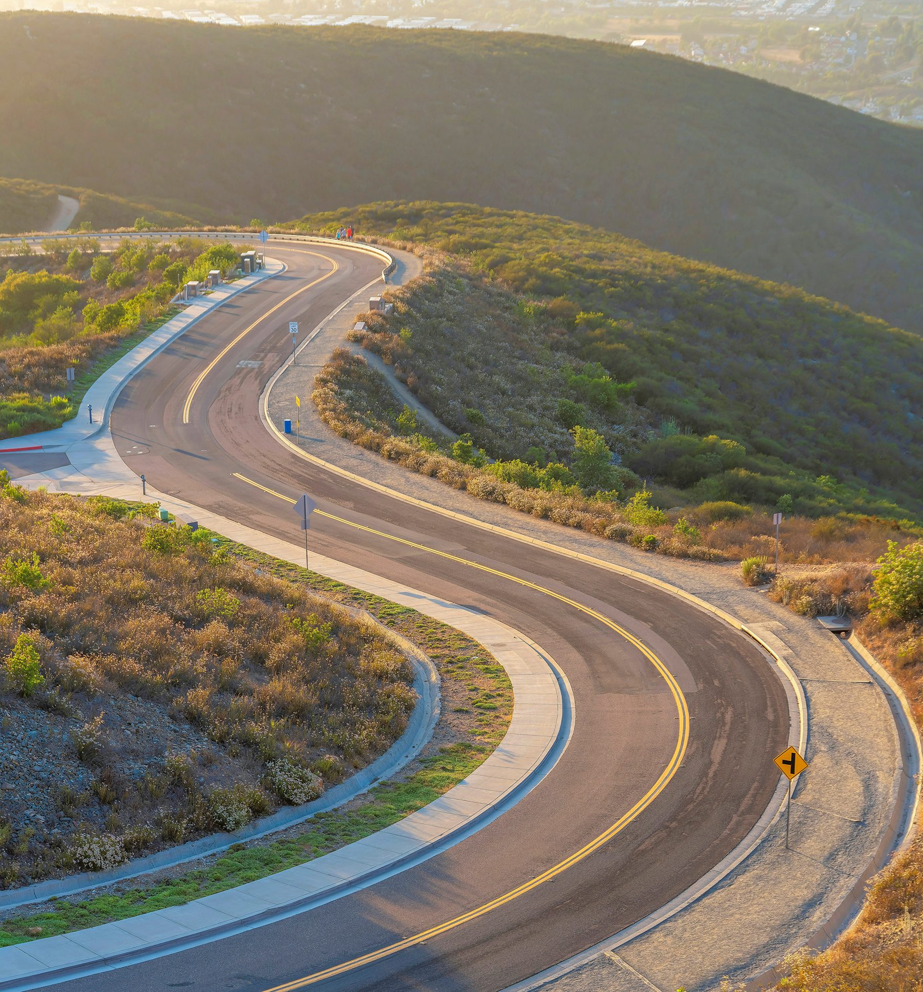 High angle view of a winding road on top of a mountain with a view of mountains and a vast field of bushes and grass.